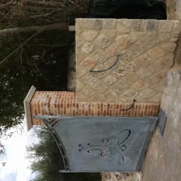 Fragment ogrodzenia z kamienia i cegły z metalową bramką z dekoracyjnym wycięciem, widoczne przewody elektryczne.