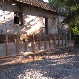 Przygotowanie do izolacji fundamentów starego domu: szalunek z desek i stalowe pręty zbrojeniowe wystające z fundamentu z kamienia, widoczne ślady zawilgocenia na ścianie budynku.