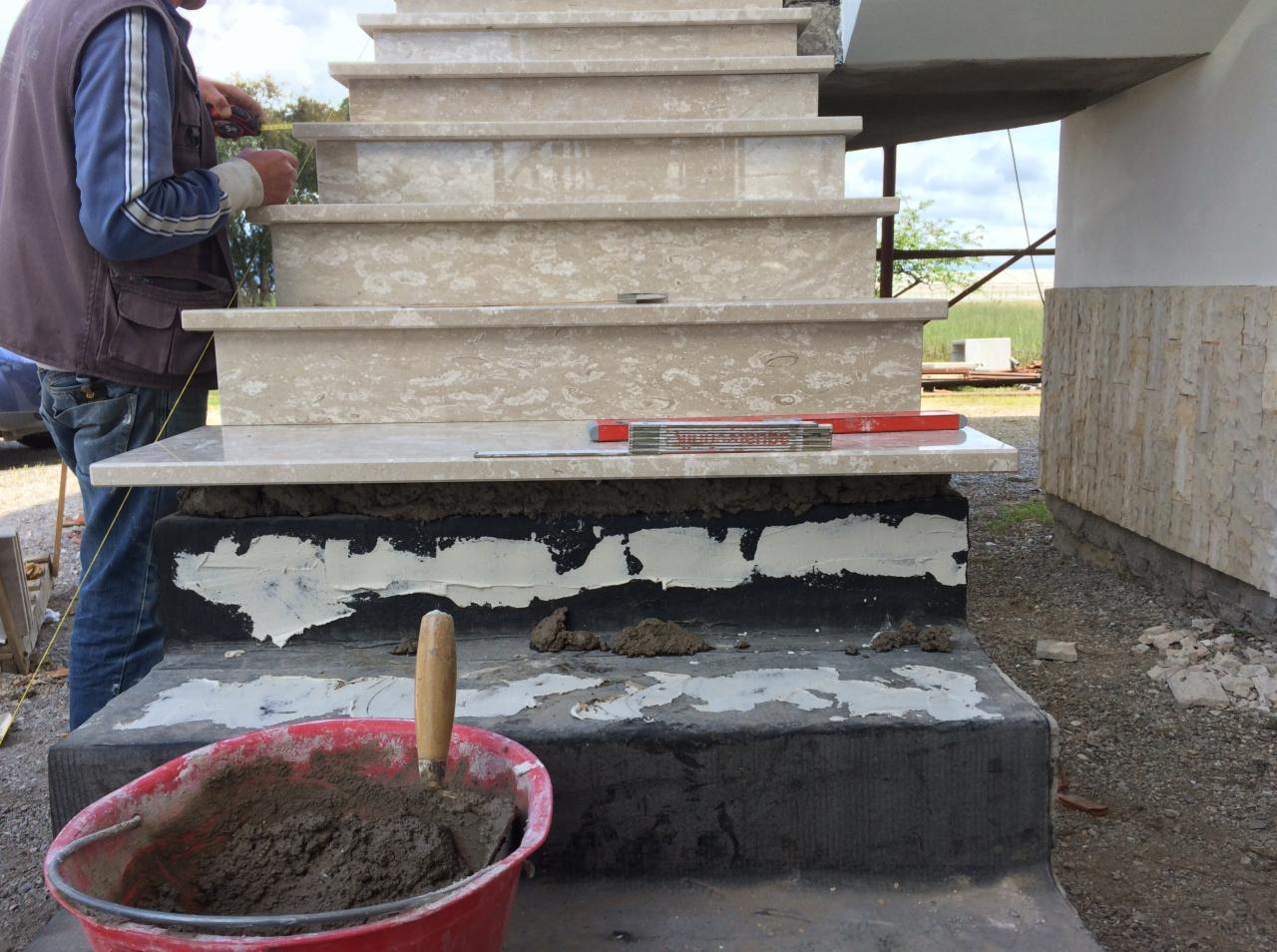 Montaż marmurowych stopni schodowych zewnętrznych, widoczny murarz z miarką, poziomica, zaprawa w czerwonym wiadrze i szpachelka.