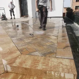 Mycie kostki brukowej na posesji z użyciem węża. Widoczny fragment posesji z ogrodzeniem i budynkiem. Prace brukarskie w toku, etap czyszczenia.