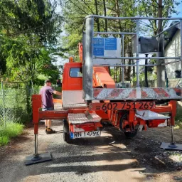 Pomarańczowy podnośnik koszowy na stabilizatorach, przygotowany do pracy w ogrodzie, w tle drzewa i siatka ogrodzeniowa, widoczny pracownik. Numer telefonu na pojeździe.