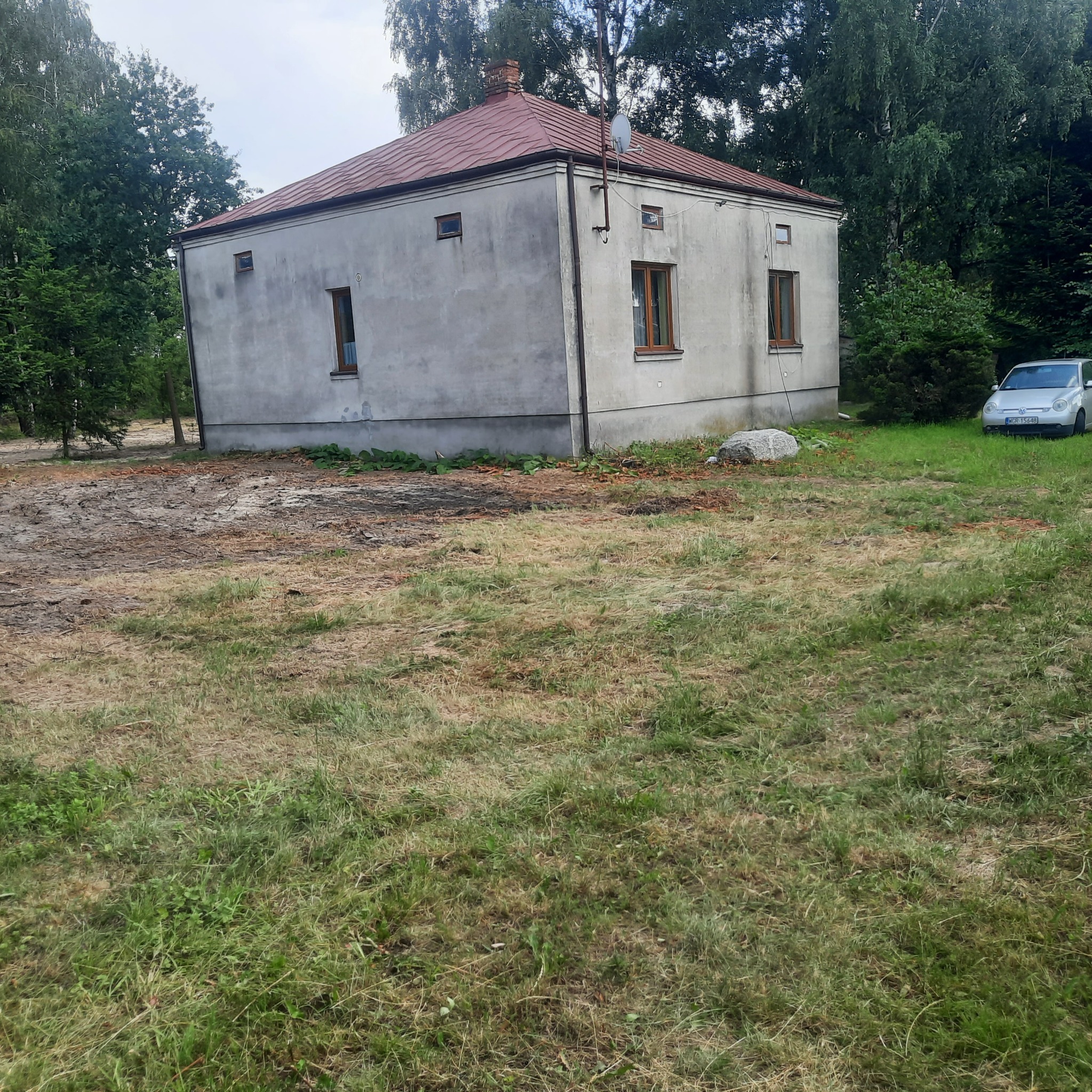 Szary, kwadratowy budynek z czerwoną dachówką, otoczony trawnikiem i drzewami, po niedawnych pracach ziemnych, z zaparkowanym samochodem w tle.