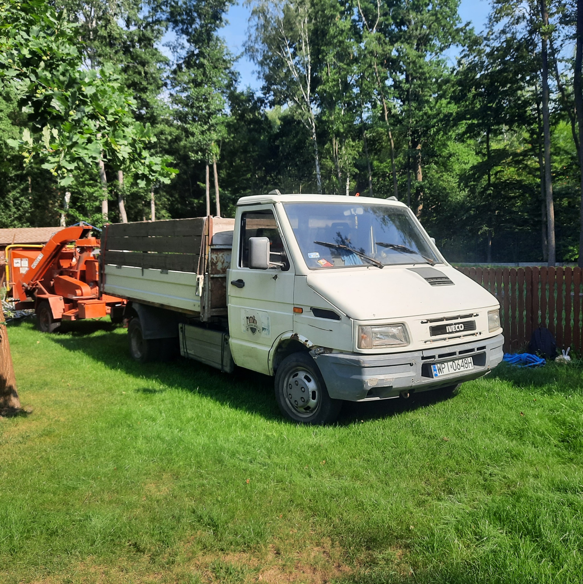 Biały samochód dostawczy Iveco z przyczepioną pomarańczową rębarką do gałęzi na trawniku, w tle drzewa i brązowe ogrodzenie.