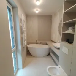 Nowoczesna łazienka w beżowej tonacji: wolnostojąca wanna, podwieszana toaleta, grzejnik drabinkowy i szafka z półkami.