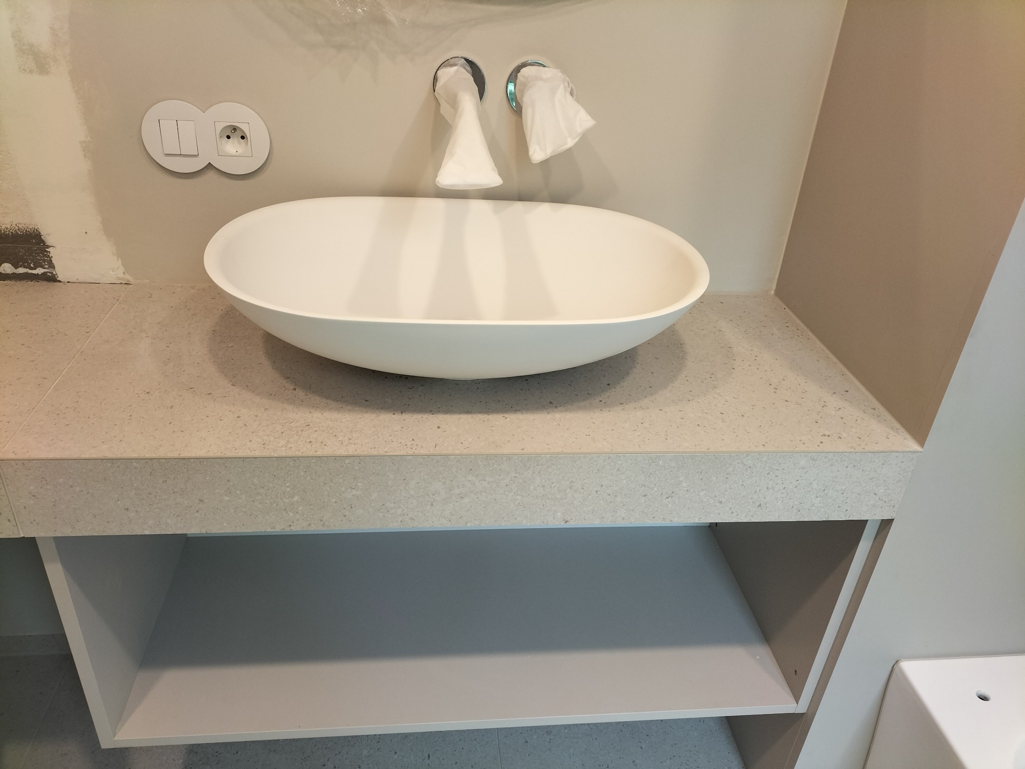 Nowoczesna, owalna umywalka nablatowa w jasnej łazience z widocznymi elementami instalacji i niedokończonym fragmentem ściany. Minimalistyczny design.