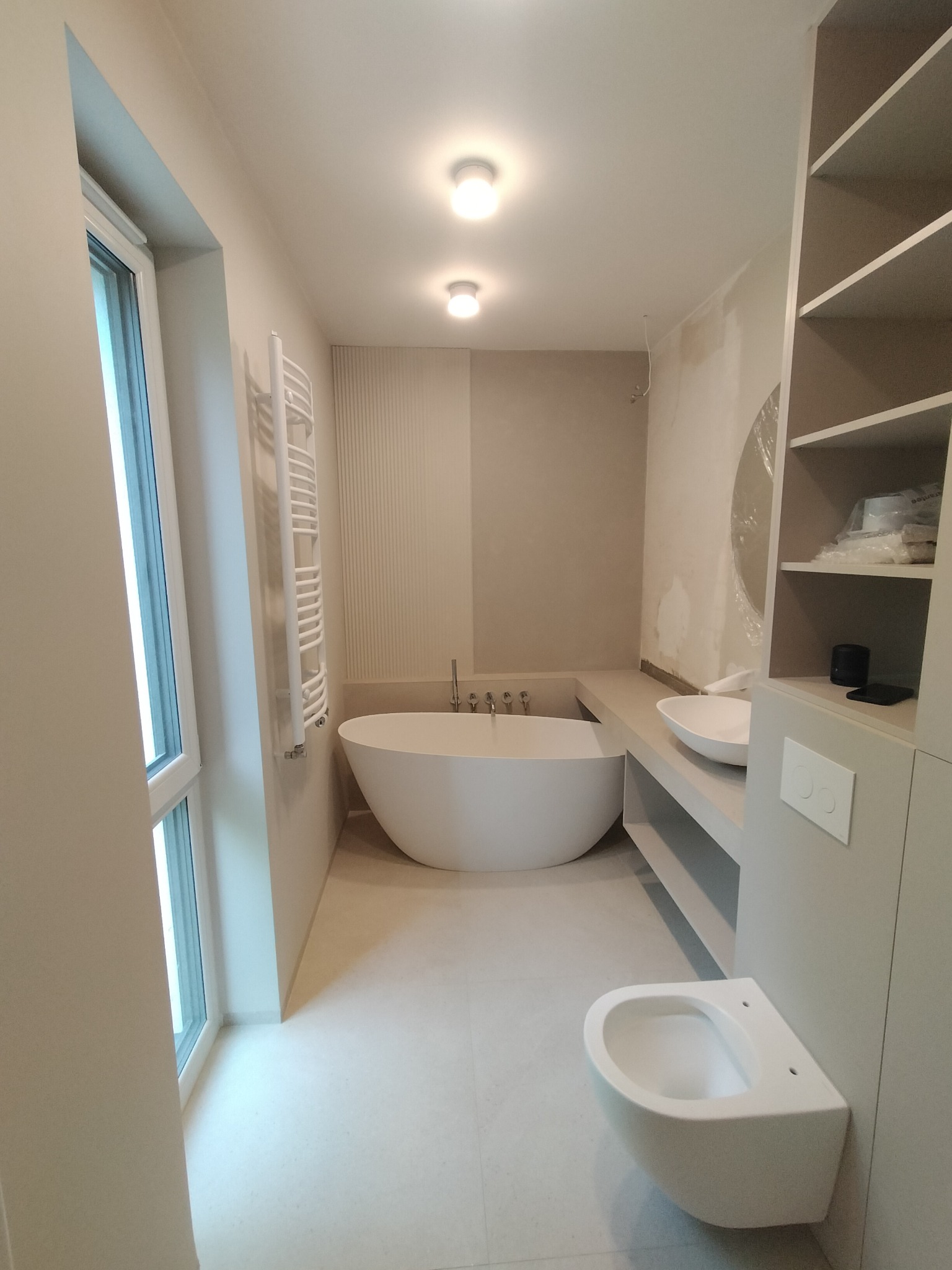 Nowoczesna łazienka w beżowej tonacji: wolnostojąca wanna, podwieszana toaleta, grzejnik drabinkowy i szafka z półkami.