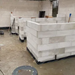 Budowa ścian domu z bloczków betonu komórkowego, widoczne spoiny i czarny kubeł na pierwszym planie, mokra posadzka, pochmurne niebo w tle.