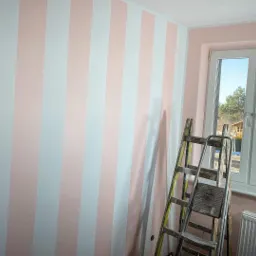 Ściana w pokoju pomalowana w pionowe pasy w kolorach białym i pudrowego różu, drabina oparta o ścianę, widoczne okno z widokiem na zewnątrz i lampa oświetleniowa