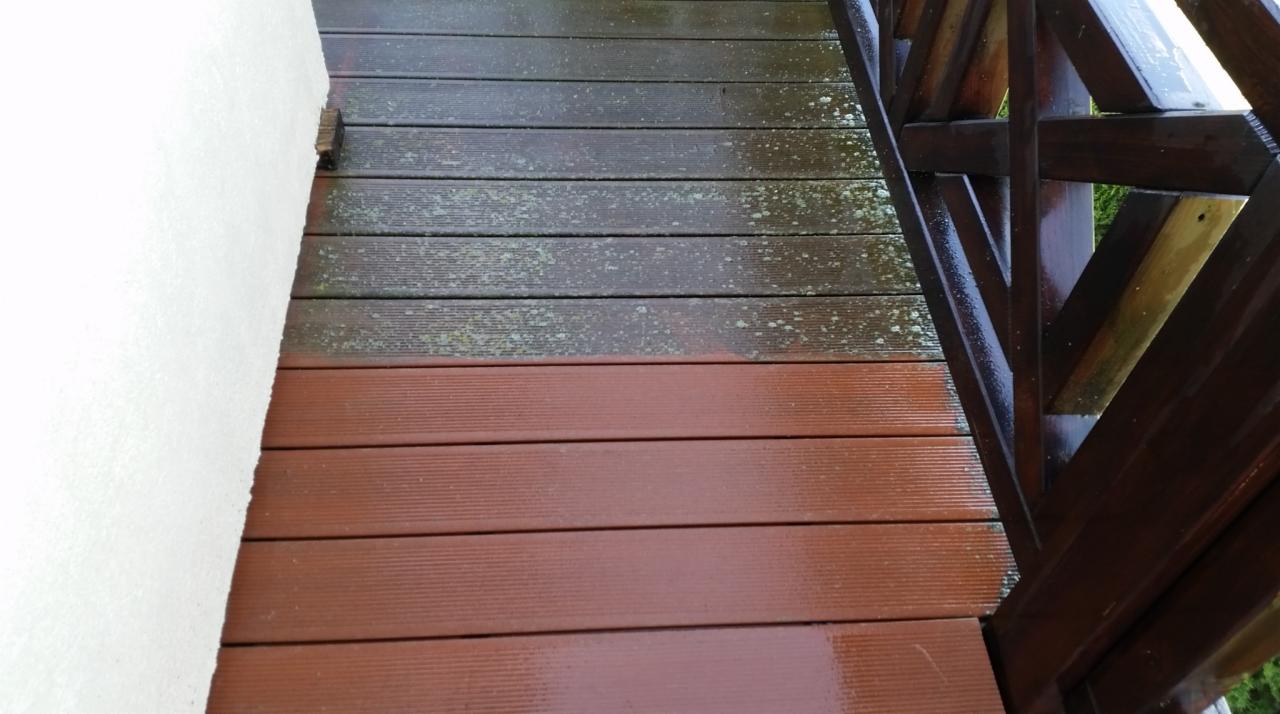 Fragment drewnianego tarasu z widoczną różnicą w czystości desek: górna część pokryta zielonym nalotem, dolna część świeżo umyta, w tle fragment ściany budynku i konstrukcja drewnianej balustrady.