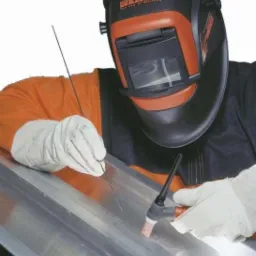 Spawacz w masce Kemppi spawa metalowy element metodą TIG, widoczne elektrody i iskry. Pracownik w kombinezonie i rękawicach ochronnych.