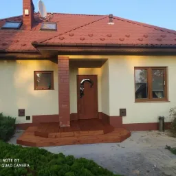 Front domu z niskim drewnianym podestem wejściowym, ceglaną kolumną i brązowymi oknami pod czerwoną dachówką.