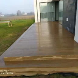 Nowoczesny, brązowy podest przy domu z wyjściem na trawnik, widok na pole w tle i stół z krzesłami w oddali, wykonany z desek kompozytowych.