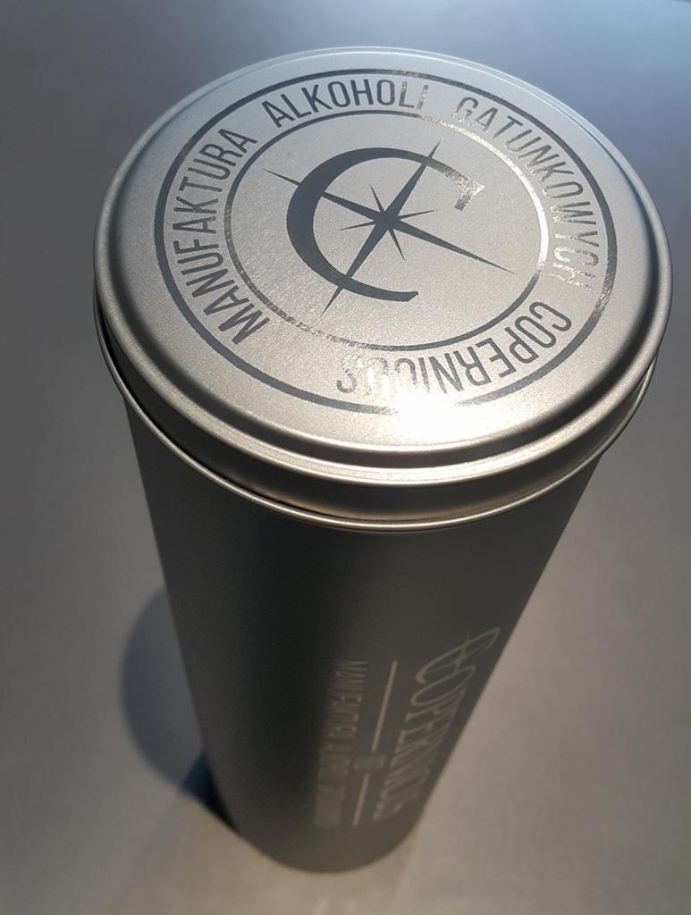 Metalowa tuba z nadrukowanym logo 'Manufaktura Alkoholi Gatunkowych Copernicus' i symbolem kompasu, widoczna z góry pod kątem.