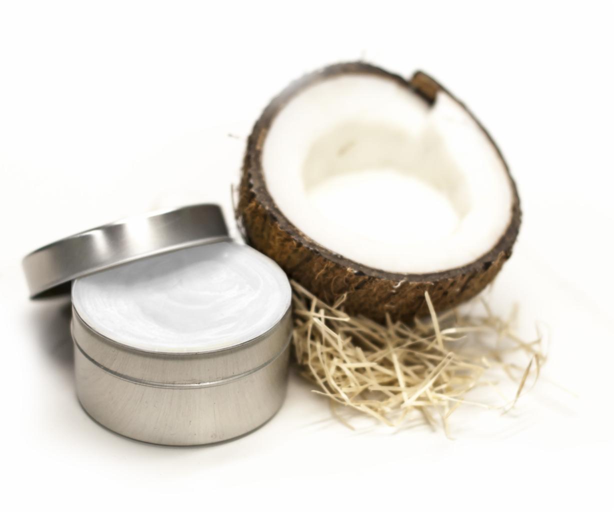 Metalowa puszka z kremem obok połówki kokosa na białym tle, z dekoracyjnym siankiem.