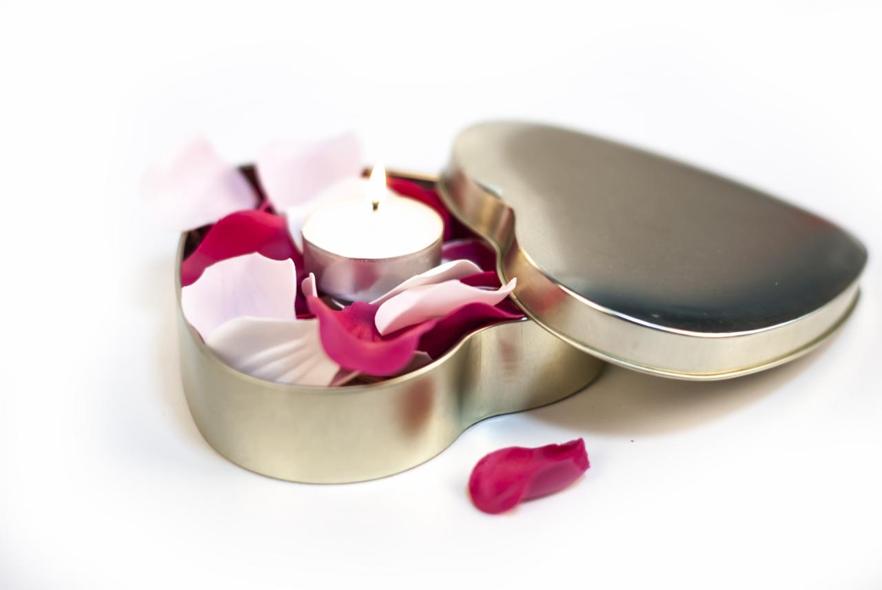 Otwarta metalowa puszka w kształcie serca wypełniona płatkami róż w odcieniach bieli i różu, z zapaloną świeczką tealight w środku, na białym tle.