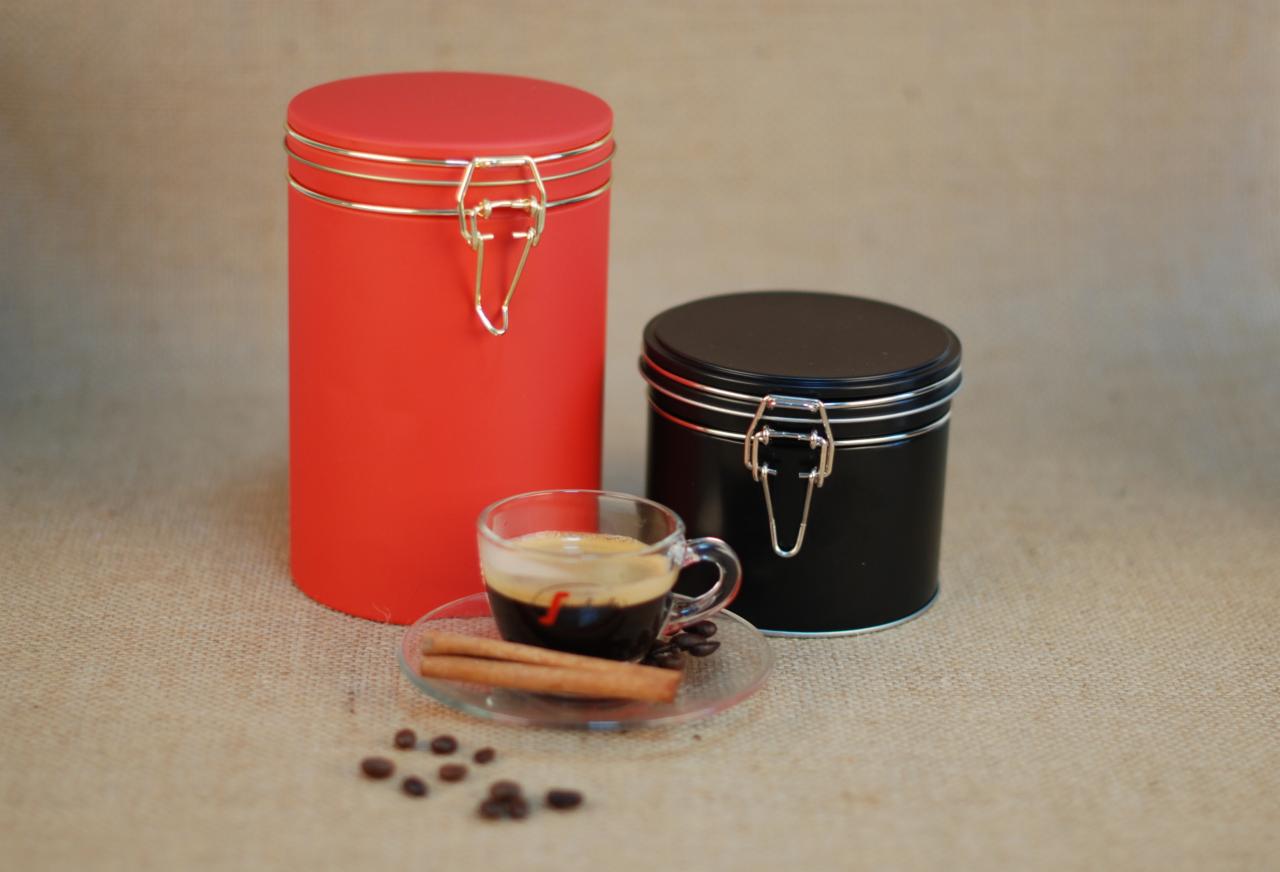 Dwa metalowe pojemniki z zamknięciem na klamrę: czerwony wysoki i czarny niski, obok filiżanka espresso z dwoma laskami cynamonu i kilkoma ziarnami kawy na tle jutowym.