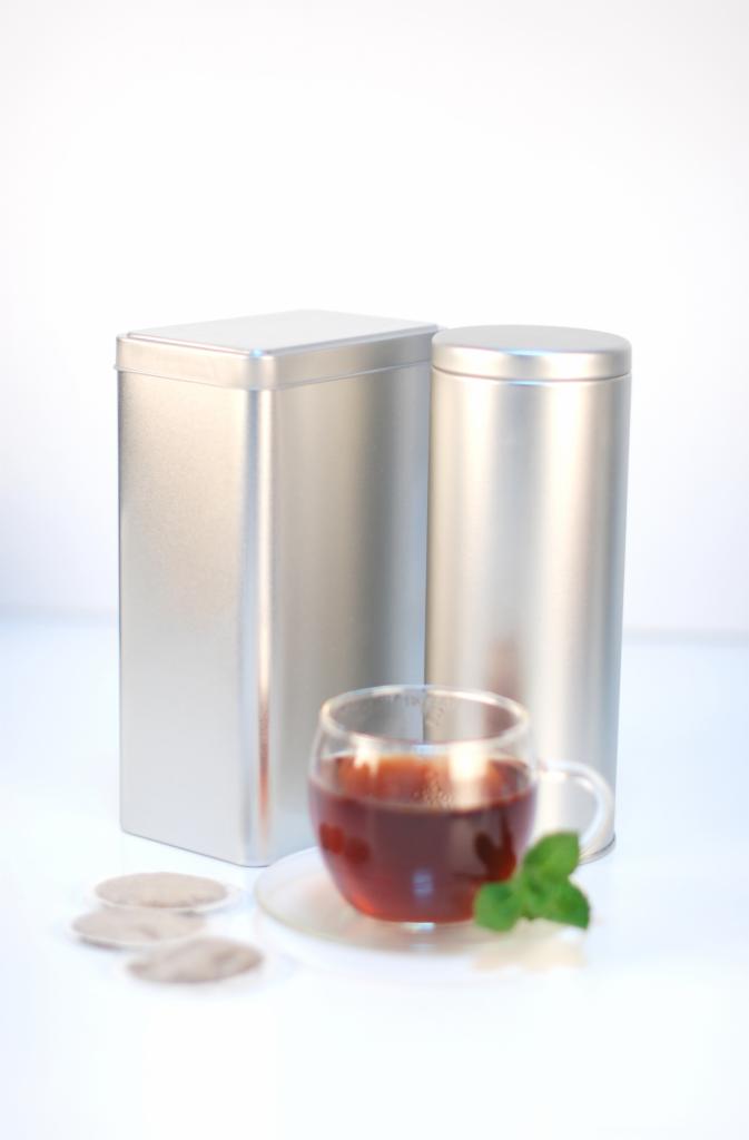 Dwie srebrne metalowe puszki, jedna kwadratowa, druga okrągła, stoją obok szklanki herbaty z listkiem mięty i trzema saszetkami herbaty na białym tle.