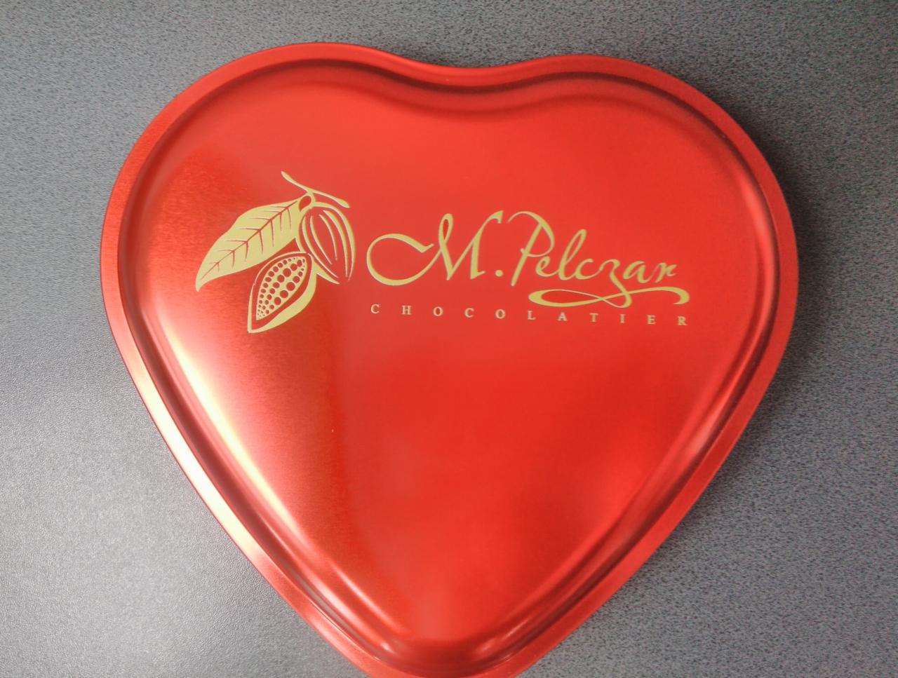 Czerwone, metalowe pudełko w kształcie serca z złotym logo i napisem 'M. Pelczar CHOCOLATIER', widoczne na szarym tle.