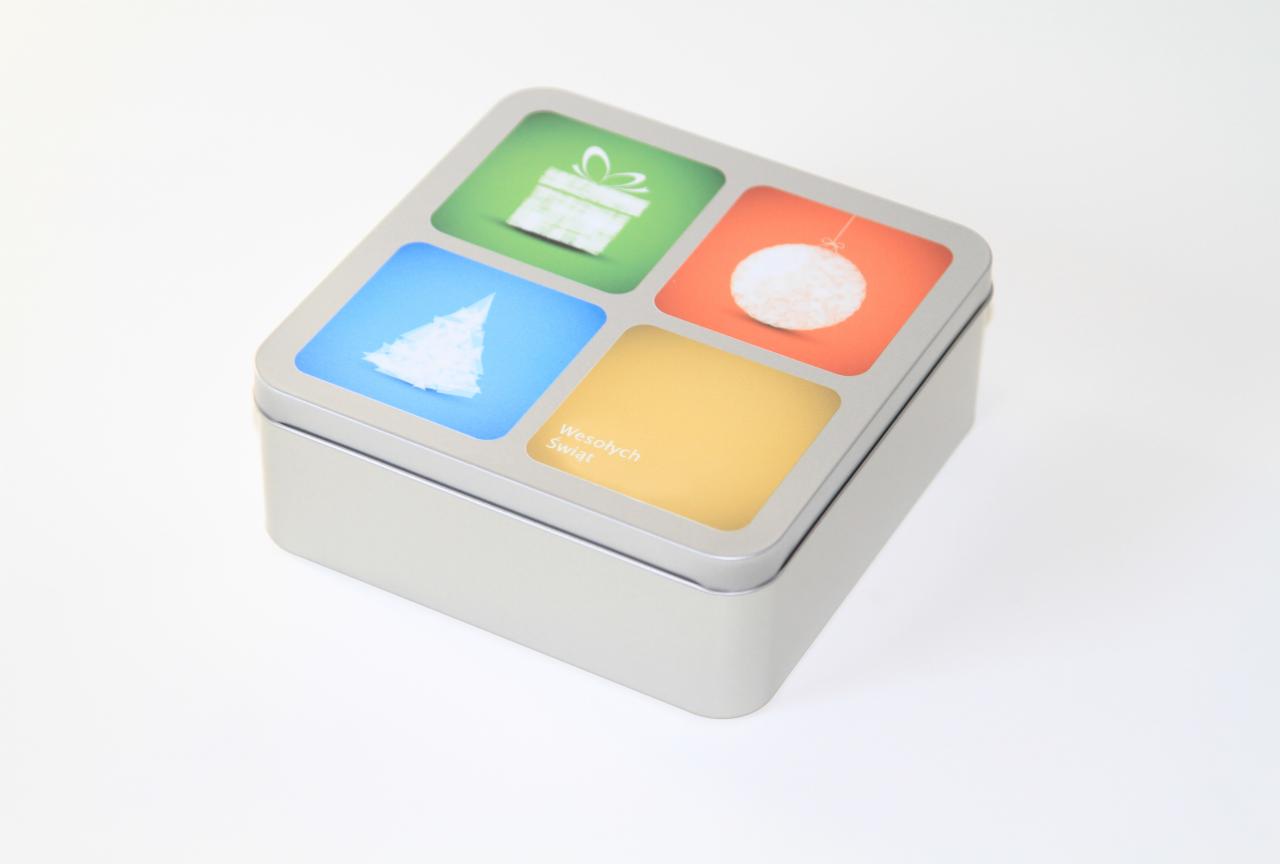 Metalowe pudełko kwadratowe z czterema kolorowymi polami na wieczku: zielone z prezentem, niebieskie z choinką, pomarańczowe z bombką i żółte z napisem 'Wesołych Świąt'.