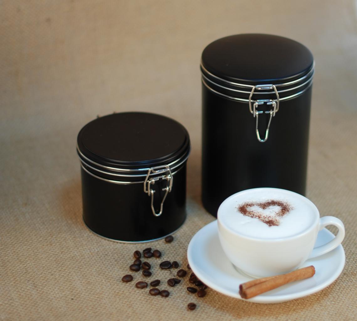 Dwie czarne metalowe puszki z zapięciem na beżowym tle, obok filiżanka cappuccino z sercem z kakao i laską cynamonu, rozrzucone ziarna kawy.