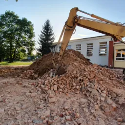 Żółta koparka nabiera gruz ceglany na tle budynku z białymi oknami i zieleni drzew