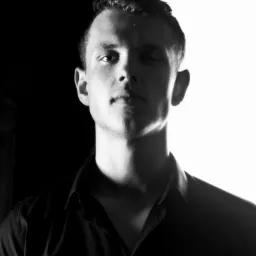 Czarno-biały portret młodego mężczyzny w ciemnej koszuli na tle podzielonym na czarną i białą część, oświetlony z góry.