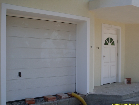 Biała segmentowa brama garażowa i drzwi wejściowe z półokrągłym oknem w jasnej elewacji budynku, widoczne cegły i żółty wąż na podłożu.