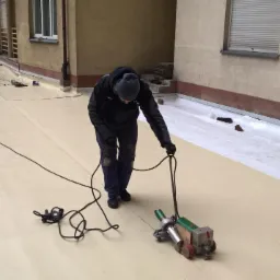 Pracownik w ciemnej odzieży używa zgrzewarki do papy termozgrzewalnej na płaskim dachu budynku z oknami i roletami.