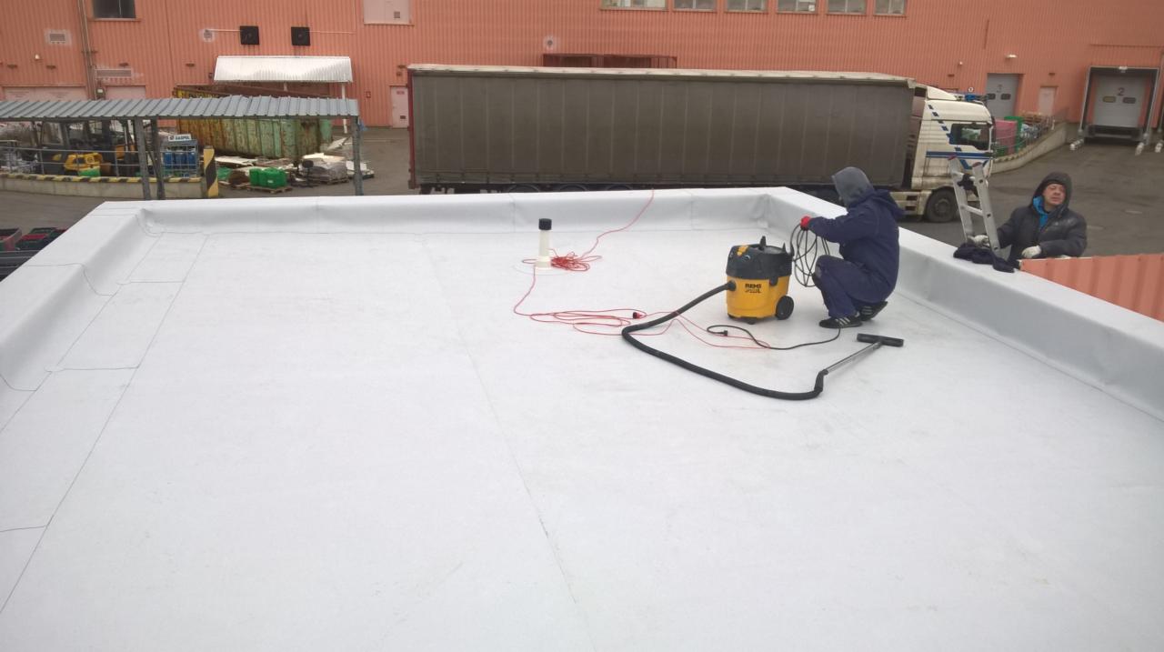Pracownik w kombinezonie czyści białą, płaską powierzchnię dachu przemysłowego za pomocą żółtego odkurzacza przemysłowego; w tle widoczny budynek przemysłowy, zaparkowana ciężarówka i drabina...