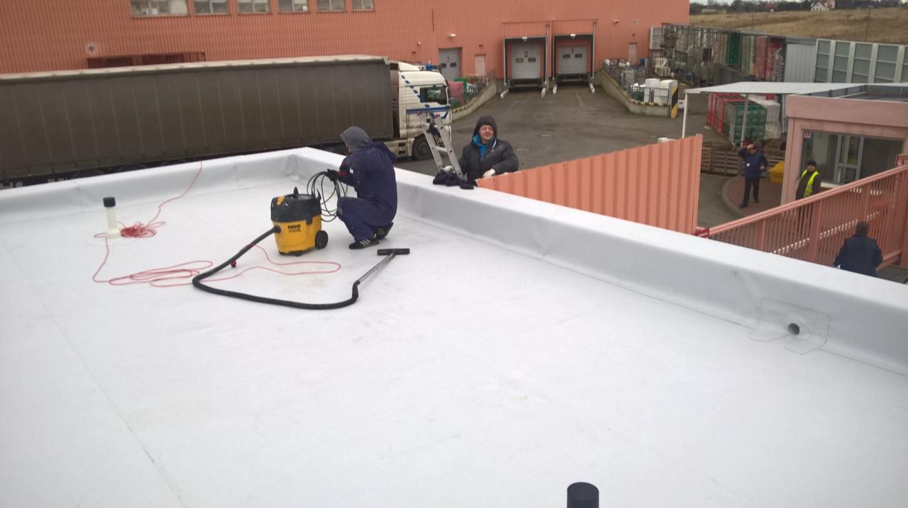 Pracownik w kombinezonie czyszczący białą powierzchnię dachu przemysłowego za pomocą odkurzacza przemysłowego, drugi pracownik obserwuje z drabiny, w tle budynki przemysłowe i zaparkowane ciężarówki.