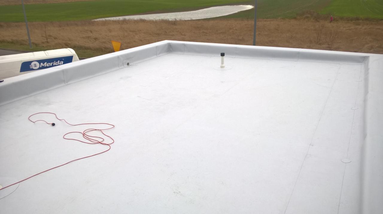 Biały dach płaski z wykończeniem, widoczna instalacja wentylacyjna, kabel na dachu, w tle budynek firmy Merida i krajobraz.