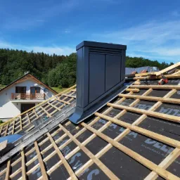 Montaż konstrukcji dachu z drewnianą więźbą, czarną membraną i kominem wentylacyjnym na tle domu i lasu, widok z góry w słoneczny dzień.