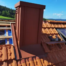 Nowy, brązowy komin z blachy na dachu z dachówki ceramicznej, widoczne narzędzia i drabinka dekarska, w tle zielone wzgórza i błękitne niebo.