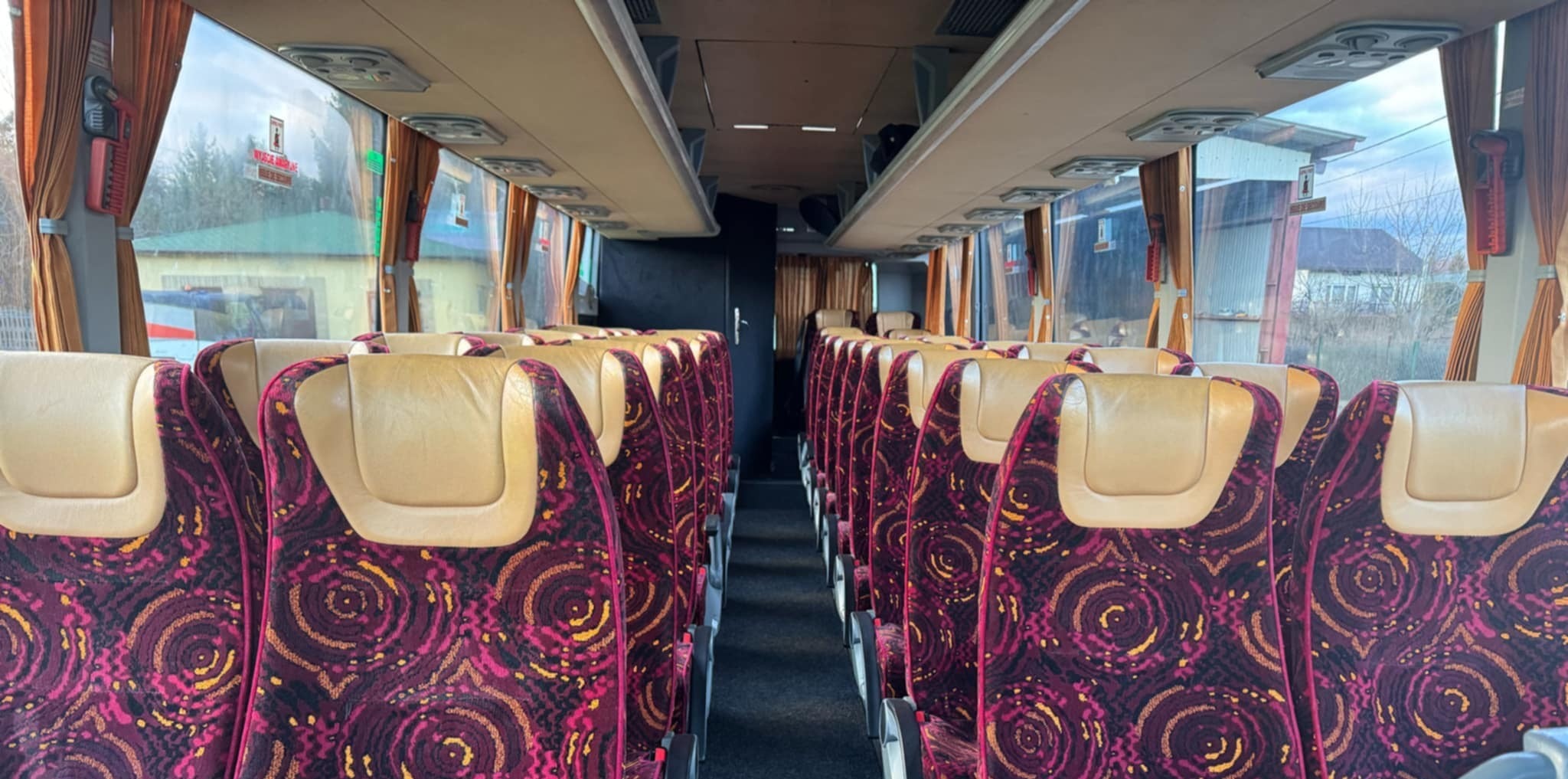 Wnętrze autobusu z beżowymi zagłówkami i bordowo-złotą tapicerką foteli. Widok na rząd siedzeń z perspektywy środka pojazdu, okna z zasłonami.