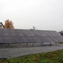 Rząd paneli słonecznych na metalowej konstrukcji, rozłożony na czarnej macie, w tle dom z ceglanym kominem i drzewo o pomarańczowych liściach pod pochmurnym niebem.