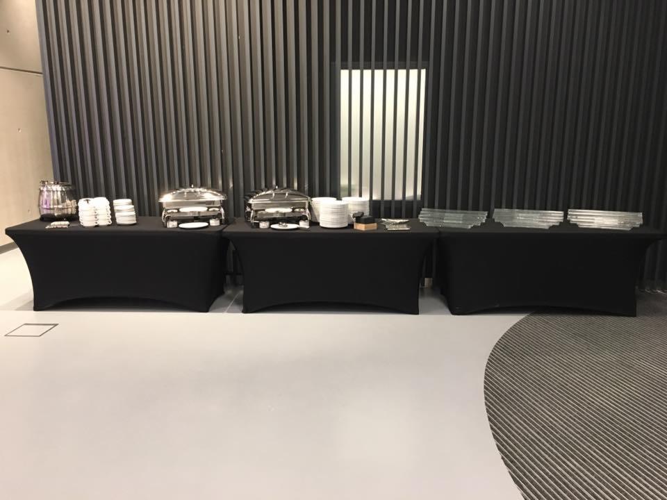 Trzy czarne stoły cateringowe z podgrzewaczami, stosem talerzy i dystrybutorem napojów ustawione na szarym tle ściany z pionowych listew.