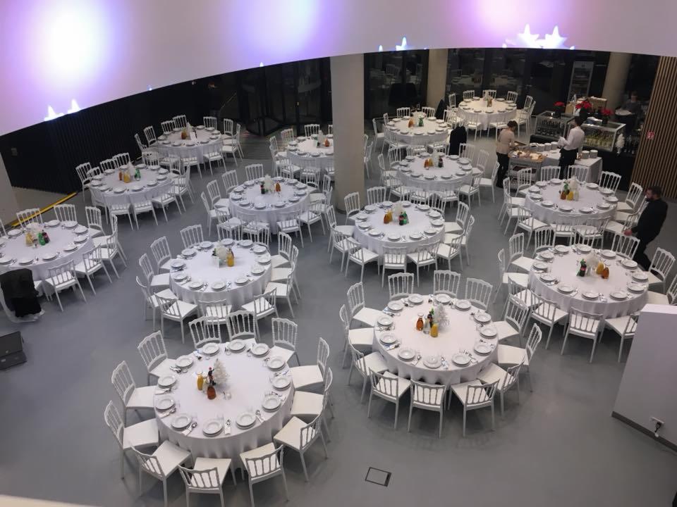 Sala bankietowa z okrągłymi stołami nakrytymi białymi obrusami i zastawą, białe krzesła ustawione wokół stołów, w tle obsługa przygotowuje posiłki.