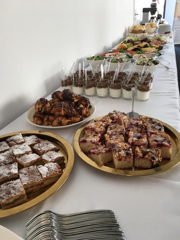 Długi stół z białym obrusem zastawiony różnorodnymi przekąskami: mini desery w szklankach z łyżeczkami, czekoladowe rogaliki, ciasto z kruszonką na złotej tacy, jabłecznik posypany cukrem pudrem...