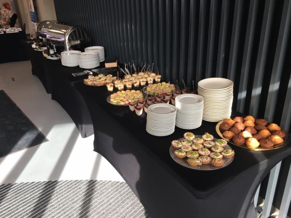 Długi stół z czarnym obrusem, na którym znajdują się talerze, mini babeczki, kanapeczki, desery w kieliszkach, makaroniki i podgrzewacze do potraw, ustawione w rzędzie wzdłuż ściany z pionowych...