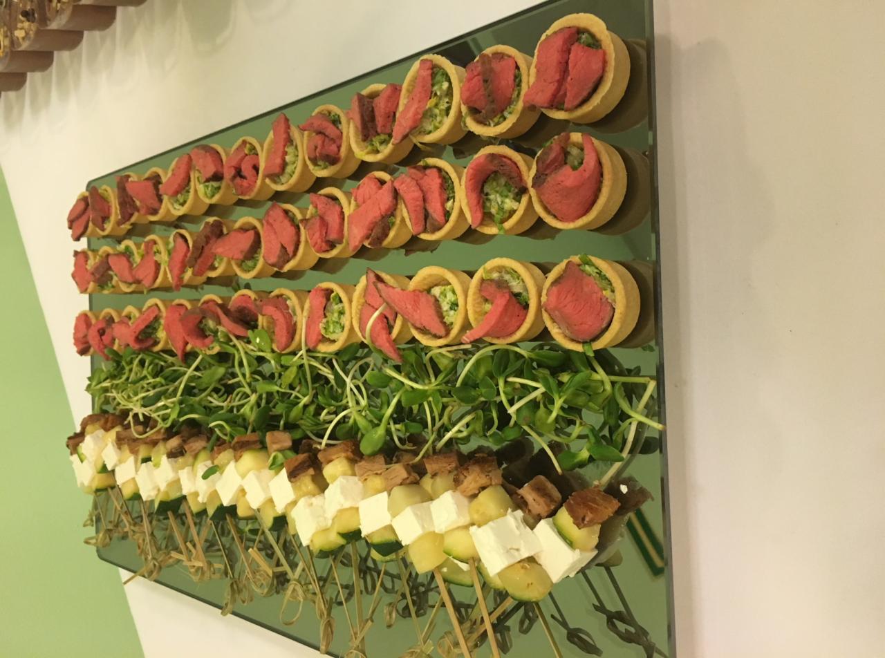 Elegancko zaaranżowane przekąski cateringowe na szklanej tacy: mini tartaletki z rostbefem i zielonym farszem oraz szaszłyki z grilla z serem feta, cukinią i ananasem, udekorowane kiełkami.