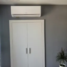 Biały klimatyzator Sinclair zamontowany nad białą szafą w pokoju z szarą ścianą, obok stoi roślina doniczkowa na drewnianym stoliku.