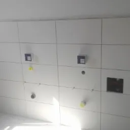 Białe płytki ceramiczne na ścianie łazienki w trakcie montażu, widoczne otwory na instalacje wodne i elektryczne, przewody i puszki pod gniazdka, poziomowanie płytek przy użyciu krzyżyków...