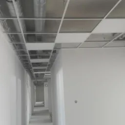 Korytarz biurowy w trakcie wykończenia, widoczne białe ściany, sufit podwieszany z odsłoniętą instalacją wentylacyjną i elektryczną.