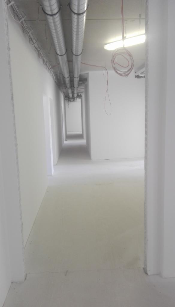 Długi, pusty korytarz w trakcie wykańczania, z widocznymi instalacjami wentylacyjnymi pod sufitem i przewodami elektrycznymi zwisającymi z sufitu.