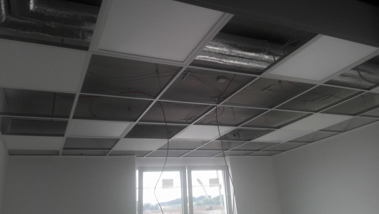 Montaż podwieszanego sufitu kasetonowego z widocznymi przewodami i instalacjami nad konstrukcją, częściowo wypełnioną białymi panelami, w nowym budynku z oknem.