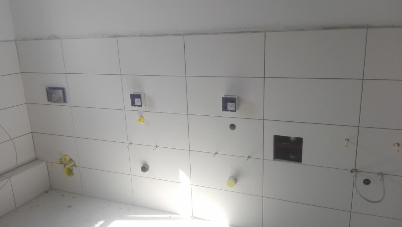 Białe płytki ceramiczne na ścianie łazienki w trakcie montażu, widoczne otwory na instalacje wodne i elektryczne, przewody i puszki pod gniazdka, poziomowanie płytek przy użyciu krzyżyków...