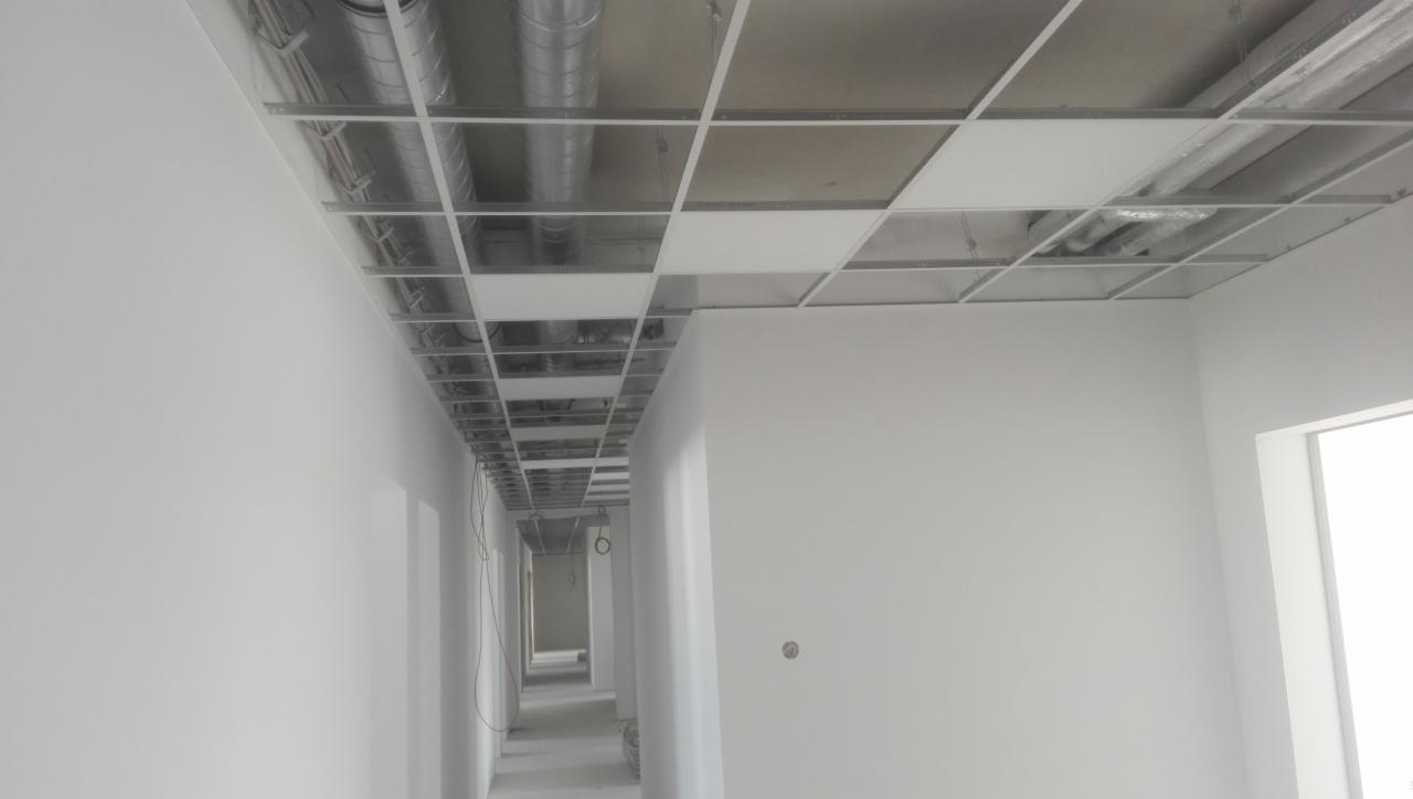 Korytarz biurowy w trakcie wykończenia, widoczne białe ściany, sufit podwieszany z odsłoniętą instalacją wentylacyjną i elektryczną.