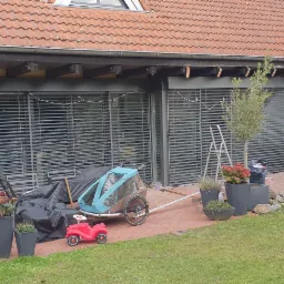 Zewnętrzne rolety aluminiowe zamontowane w oknach domu jednorodzinnego, widok z ogrodu, na pierwszym planie wózek rowerowy i zabawka dla dziecka.