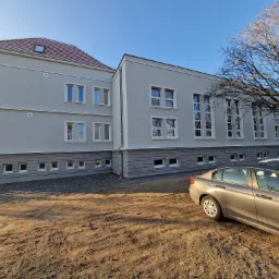 Szary budynek z białymi ramami okiennymi i brązowym dachem w słoneczny dzień, z zaparkowanym samochodem na pierwszym planie i drzewem z gołymi gałęziami po prawej stronie.