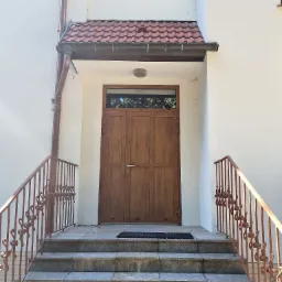 Wejście do budynku z dwuskrzydłowymi drzwiami z górnym naświetlem, zadaszone czerwonymi dachówkami, prowadzące po schodach z metalową balustradą.